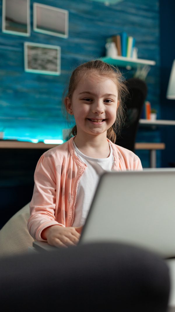 Portrait of smiling schoolkid looking into camera while studying literature for online class lesson sitting on big bean in living room. Clever little child working at mathematics homework